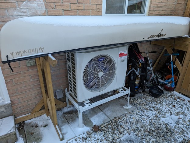 A heat pump on a stand located outside a home with a canoe stored upside down over top of it.