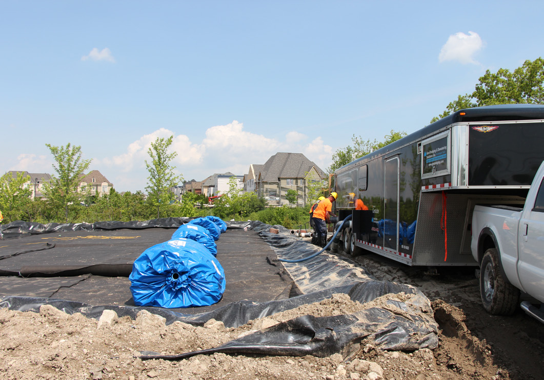 Figure 5. Sediment dewatering bag laydown area. Several large blue dewatering bags are placed on a lined surface while two workers in safety vests set up equipment beside the trailer.